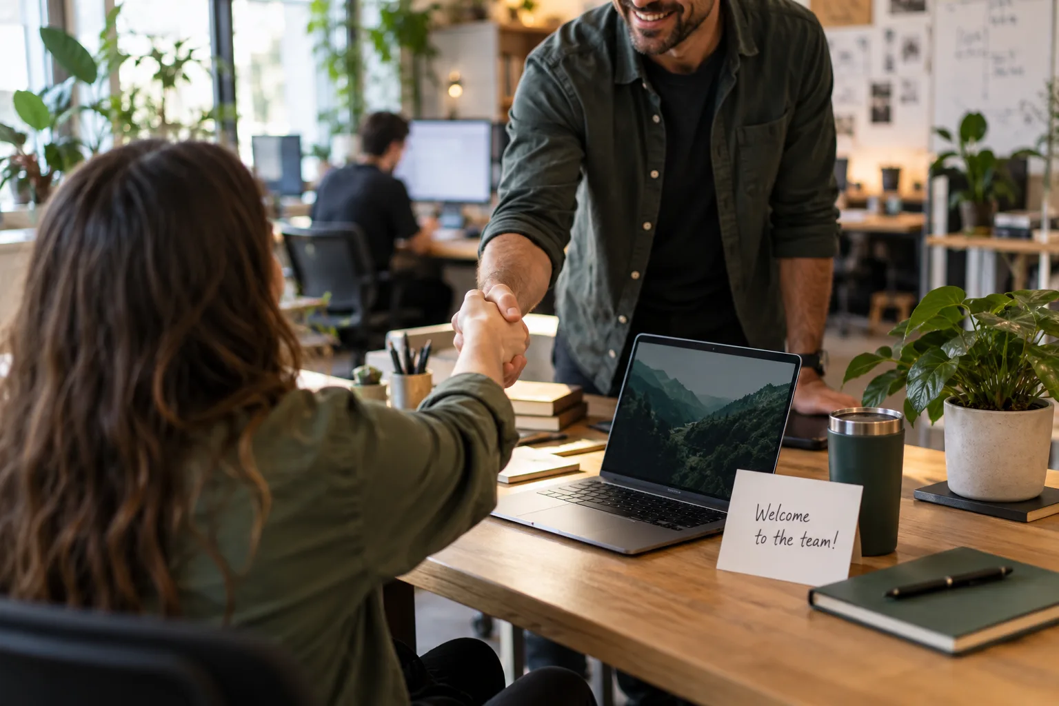 A new hire being welcomed with a handshake, a card on the desk reading welcome to the team.